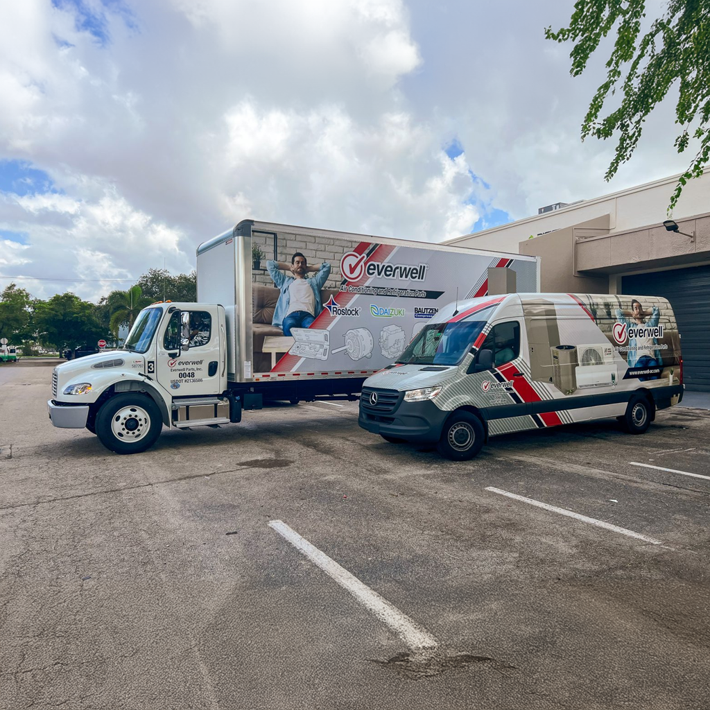 Two branded Everwell vehicles—a box truck and a cargo van—are parked side-by-side in a commercial lot, both featuring coordinated graphics with medical imagery and red-gray accents.